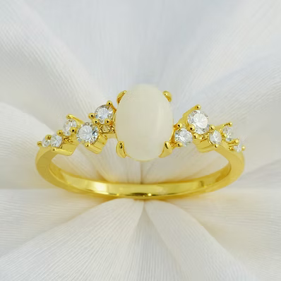 A gold ring featuring a large oval cream cabochon center stone and small clear round stones on either side, displayed on a white fabric background.