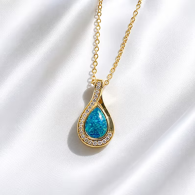 Gold teardrop pendant necklace with a blue opal center and clear crystal accents, displayed on a white satin fabric background.