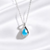 A silver teardrop pendant necklace featuring a vibrant blue teardrop gemstone surrounded by a halo of small clear crystals, displayed on a soft white satin fabric background.