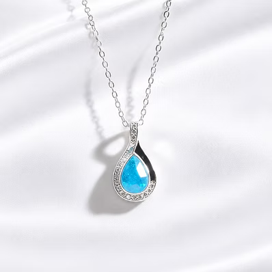 A silver teardrop pendant necklace featuring a vibrant blue teardrop gemstone surrounded by a halo of small clear crystals, displayed on a soft white satin fabric background.