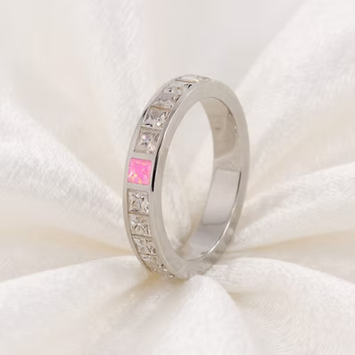 A silver band ring featuring a row of clear square-cut stones and a central pink stone, displayed on a soft white fabric background.