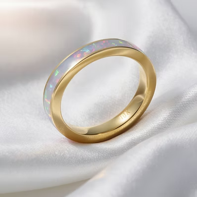 A gold band ring featuring a smooth, iridescent white opal inlay with subtle pink and blue flashes, resting on a draped white satin fabric.