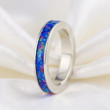 A silver band ring featuring a vibrant, multicolored inlay of blue, purple, and orange opal, displayed on a soft white satin fabric background.