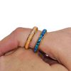 Two gold band rings worn on a person's fingers, one featuring a warm orange-pink iridescent inlay and the other a vivid blue-purple iridescent inlay, against a plain white background.