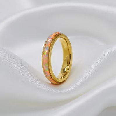 A gold band ring featuring a vibrant iridescent inlay of pink, orange, and blue opal, resting on a draped white satin fabric.