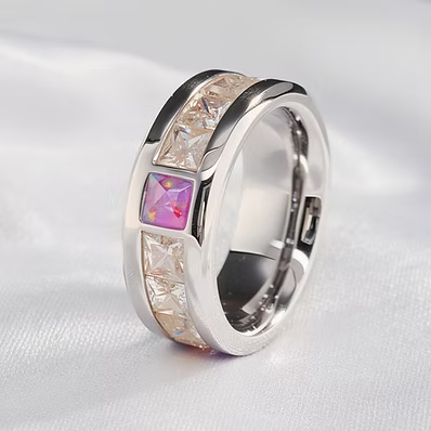 A silver-toned ring with a polished band, featuring a central square pink gemstone and clear crystal-like accents on a soft white fabric background.