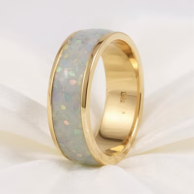 A polished yellow gold ring featuring a smooth, inlaid white opal band with iridescent flecks, displayed on a soft white fabric background.