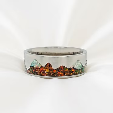 A silver-toned band ring featuring an inlaid mountain landscape design with green, orange, and brown stones, set against a white background.
