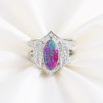 A silver ring featuring a large marquise-cut iridescent opal center stone with wing-like side details and small clear stones, displayed on a soft white fabric background.