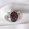 A silver ring with a wing-shaped band and a central oval cabochon stone showing a multicolored iridescent opal-like pattern, displayed on a soft white satin fabric.