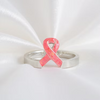 A silver ring featuring a pink opal awareness ribbon charm, displayed on a white satin fabric background.