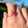 A close-up of a hand wearing a silver ring shaped like a pink awareness ribbon, photographed outdoors with a blurred natural background.