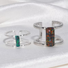 Two silver-toned open cuff rings displayed on a white satin fabric background. One ring features a vertical rectangular green opal inlay, and the other features a larger vertical rectangular multicolor opal inlay.