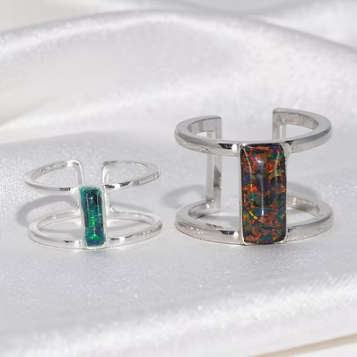 Two silver-toned open cuff rings displayed on a white satin fabric background. One ring features a vertical rectangular green opal inlay, and the other features a larger vertical rectangular multicolor opal inlay.