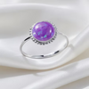 A silver ring featuring a round purple opal cabochon with iridescent blue flashes, set in a beaded silver halo on a polished silver band, resting on draped white satin fabric.