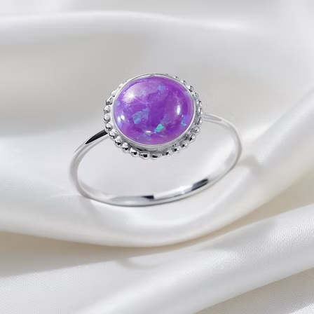 A silver ring featuring a round purple opal cabochon with iridescent blue flashes, set in a beaded silver halo on a polished silver band, resting on draped white satin fabric.