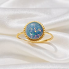 A gold ring with a round, iridescent blue opal-like center stone, resting on draped white satin fabric.