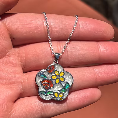 A silver pendant necklace with colorful enamel floral details, held in a person's hand.