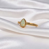 A gold ring featuring a teardrop-shaped opal gemstone with iridescent green and blue flashes, set on a textured twisted band, displayed on draped white satin fabric.