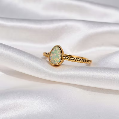 A gold ring featuring a teardrop-shaped opal gemstone with iridescent green and blue flashes, set on a textured twisted band, displayed on draped white satin fabric.