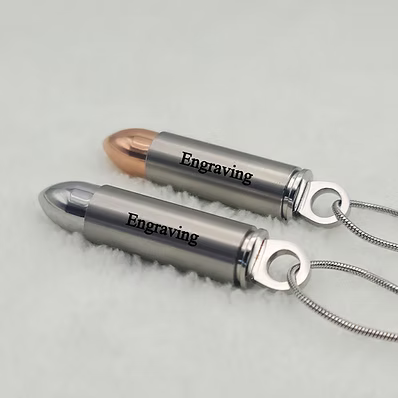 Two bullet-shaped pendant necklaces with engraved text, one in polished silver and one in rose gold, resting on a white textured surface.