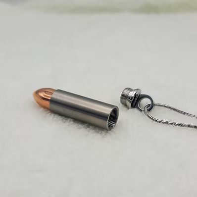A bullet-shaped pendant necklace made of silver-colored metal, shown disassembled with a copper-colored bullet tip and a silver chain attached, resting on a white textured surface.