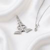 A silver pendant necklace featuring a rolled book with wings and a small heart-shaped charm on a delicate chain, displayed on a draped white satin fabric.