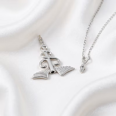 A silver pendant necklace featuring a rolled book with wings and a small heart-shaped charm on a delicate chain, displayed on a draped white satin fabric.