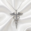 A silver caduceus pendant necklace with wings and a chain, displayed on a white satin fabric background.