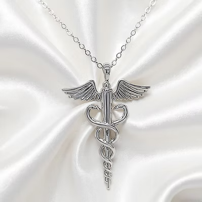 A silver caduceus pendant necklace with wings and a chain, displayed on a white satin fabric background.