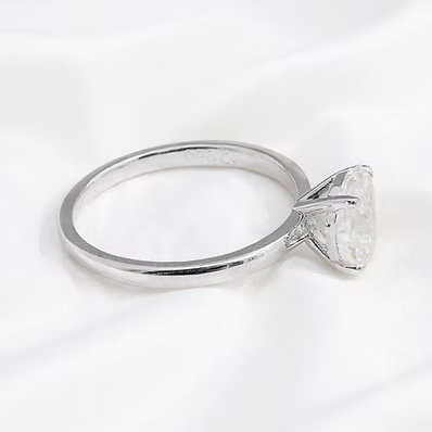 A silver-toned solitaire ring with a large clear triangular gemstone, displayed on a soft white fabric background.