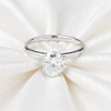 A silver solitaire engagement ring with a round brilliant-cut clear gemstone, displayed on a textured white satin fabric background.