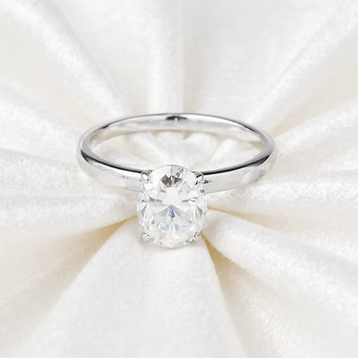 A silver solitaire engagement ring with a round brilliant-cut clear gemstone, displayed on a textured white satin fabric background.