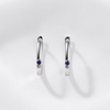 A pair of polished silver huggie hoop earrings, each featuring a small round-cut deep blue gemstone in the center, displayed on a clean white background with soft shadows.