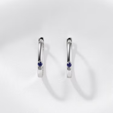 A pair of polished silver huggie hoop earrings, each featuring a small round-cut deep blue gemstone in the center, displayed on a clean white background with soft shadows.