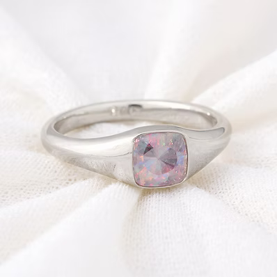 A silver band ring featuring a cushion-cut pink opal gemstone, resting on a soft, textured white fabric background.
