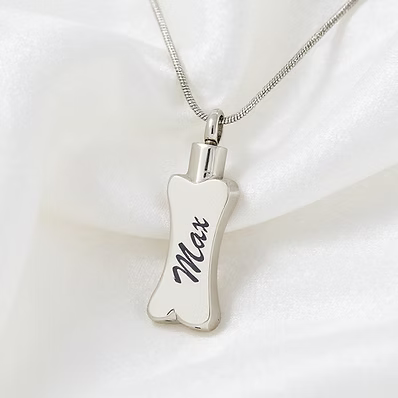 A silver bone-shaped pendant necklace engraved with the name 'Max' in black script, resting on a soft white satin fabric background.