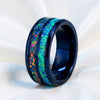 A black ceramic ring with a vibrant, multicolored iridescent inlay featuring shades of green, blue, orange, and purple, displayed on a soft white fabric background.