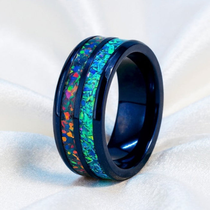 A black ceramic ring with a vibrant, multicolored iridescent inlay featuring shades of green, blue, orange, and purple, displayed on a soft white fabric background.