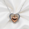 A heart-shaped pendant necklace featuring a polished rose gold heart suspended inside an open silver heart frame, displayed on a soft white satin fabric background.