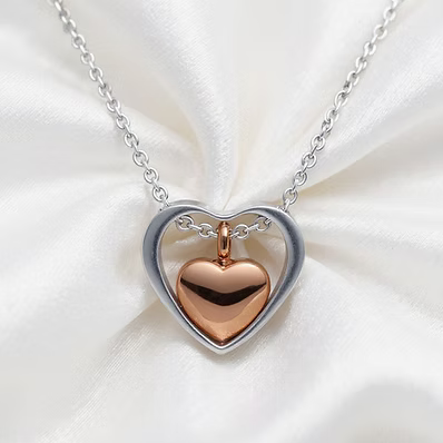 A heart-shaped pendant necklace featuring a polished rose gold heart suspended inside an open silver heart frame, displayed on a soft white satin fabric background.