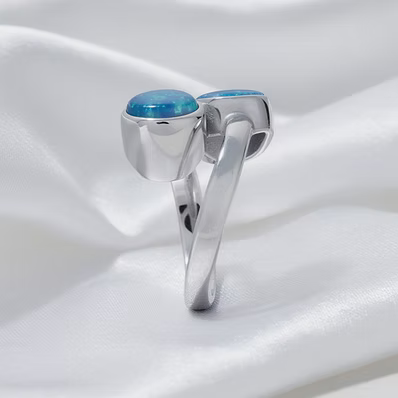 A polished silver ring featuring two oval-cut light blue gemstones, displayed on a draped white satin fabric background.