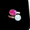 A silver-toned open ring featuring two oval cabochon gemstones, one vibrant pink and one milky white with blue iridescence, displayed on a black background.
