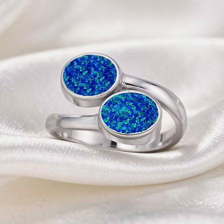 A silver ring with two round bezel-set blue glitter stones, displayed on a soft white satin fabric background.