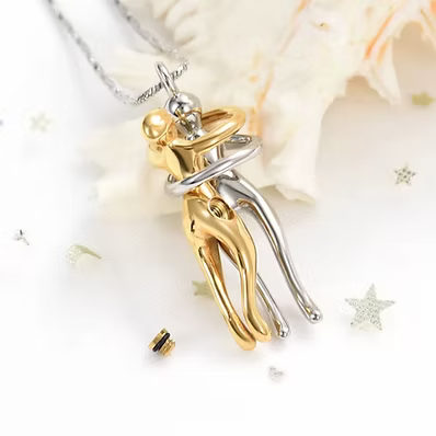 A two-tone metal pendant necklace featuring a gold and silver figurine of a couple embracing, suspended from a silver chain. The pendant is displayed on a white surface decorated with a seashell and small star-shaped embellishments.