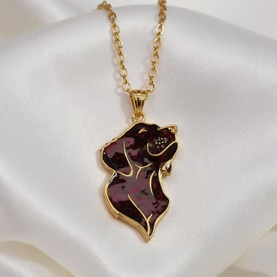 A gold chain necklace with a pendant shaped like a stylized dog head, featuring dark burgundy opal inlay with small pink flecks and gold outlines, displayed on a white satin background.