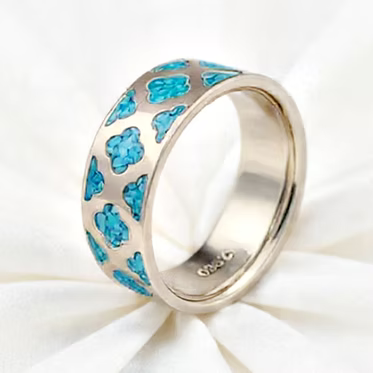 A silver band ring with a repeating pattern of bright blue clover-like shapes, displayed on a white fabric background.