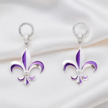 A pair of silver-tone fleur-de-lis drop earrings with purple and white enamel detailing, displayed on a draped white satin fabric background.