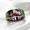 A brass-toned ring with an intricate leaf inlay design featuring green, red, and pink stones, set against a white satin background.
