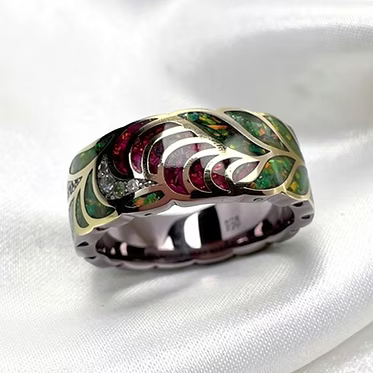 A brass-toned ring with an intricate leaf inlay design featuring green, red, and pink stones, set against a white satin background.